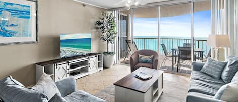 A modern living room with a large TV, comfortable seating, a ceiling fan, and a balcony with a view of the ocean.