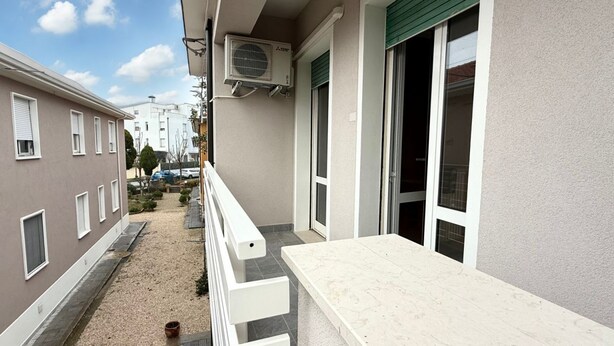 Ein Balkon mit weißer Brüstung, ein beige farbenes Gebäude mit grünem Rollo und eine Klimaanlage.