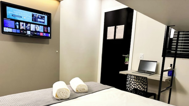 A hotel room with a bed, a desk with a laptop, a TV mounted on the wall, and a black door with a sign. - Hotel São José (Sao Jose)