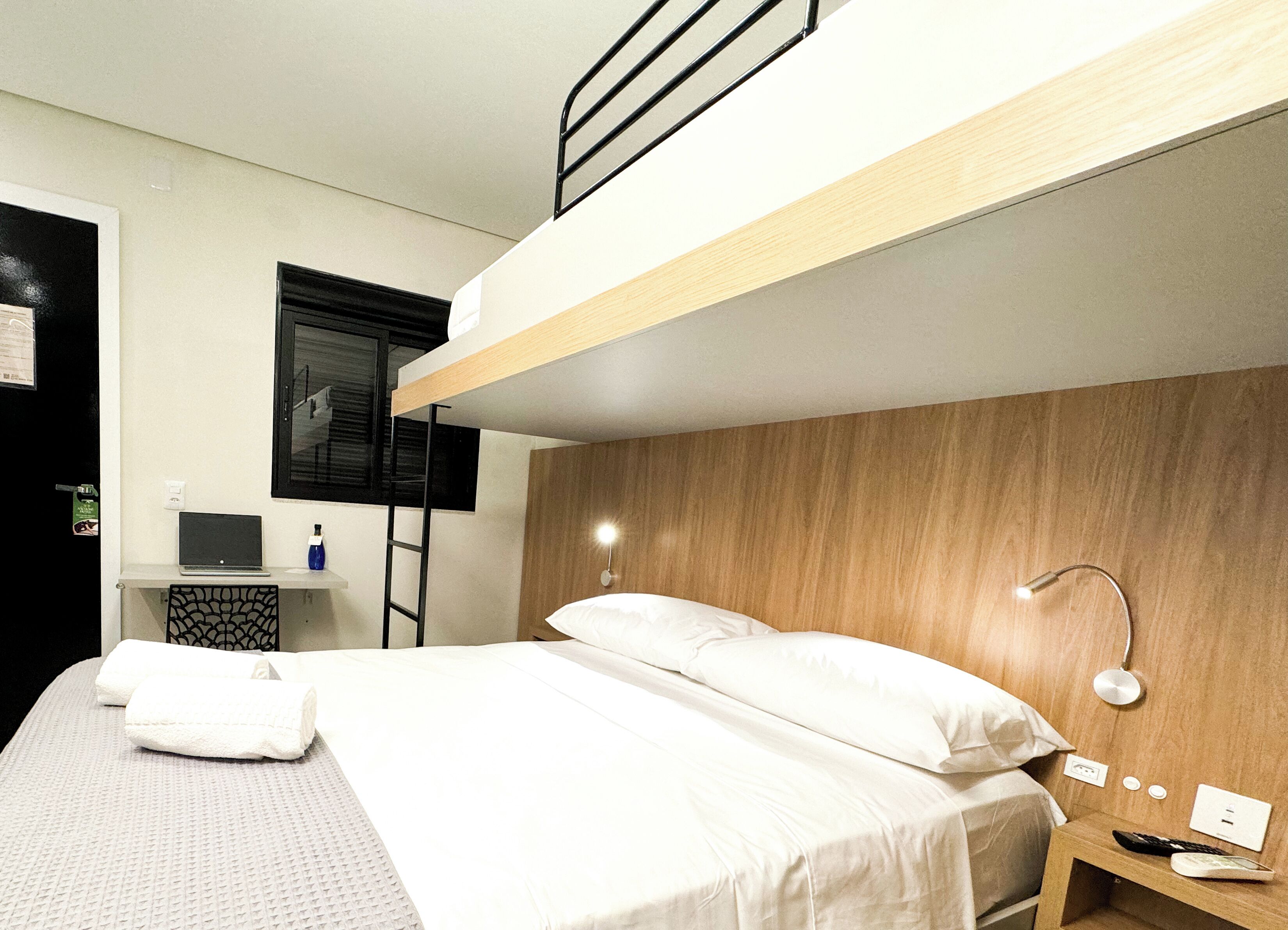 A compact hotel room with a bunk bed, a desk with a laptop, a TV, and a bedside lamp.
