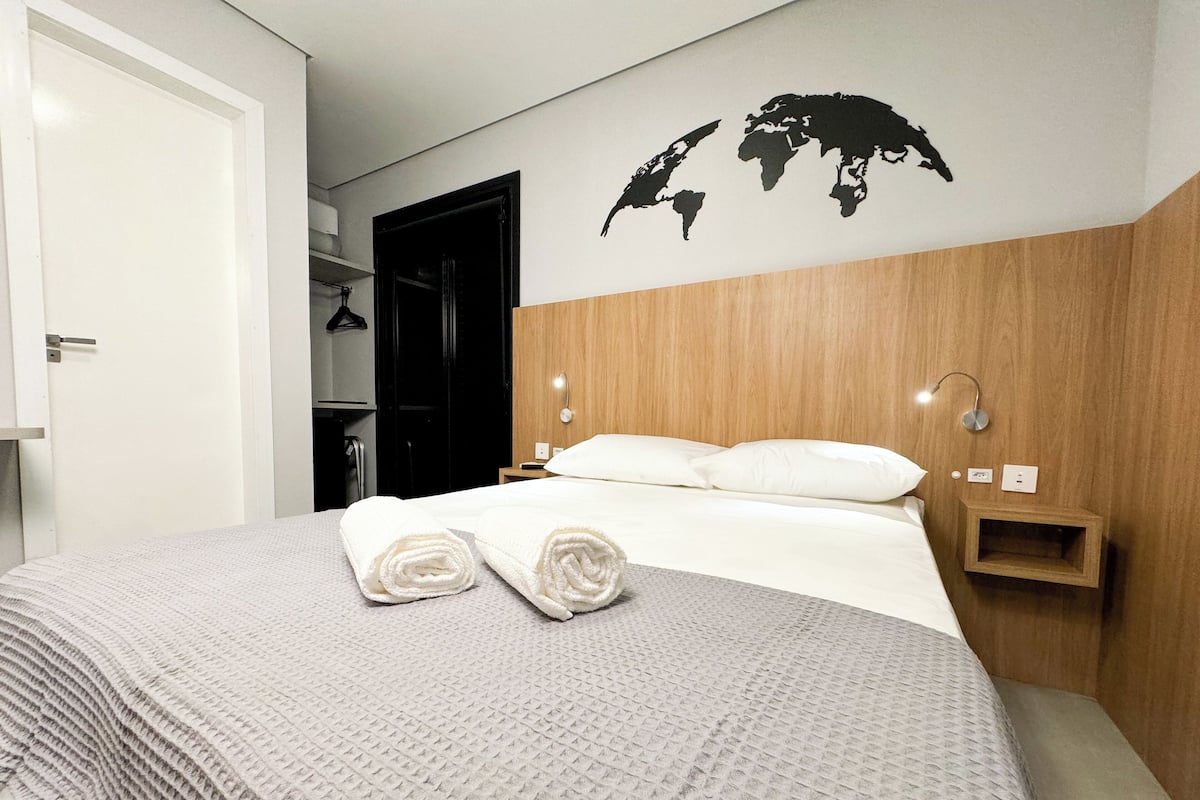 A neatly made bed with white linens and rolled towels, a wooden headboard, a world map wall decal, and a small bedside table with a lamp.