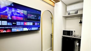 A modern bathroom with a large mirror, a flat-screen TV mounted on the wall displaying various streaming services, a microwave, and a small sink area. - Hotel São José (Sao Jose)