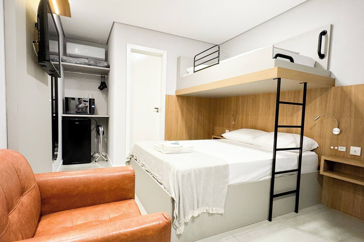 A compact room with a bunk bed, a small kitchenette, a single orange chair, and a TV mounted on the wall.