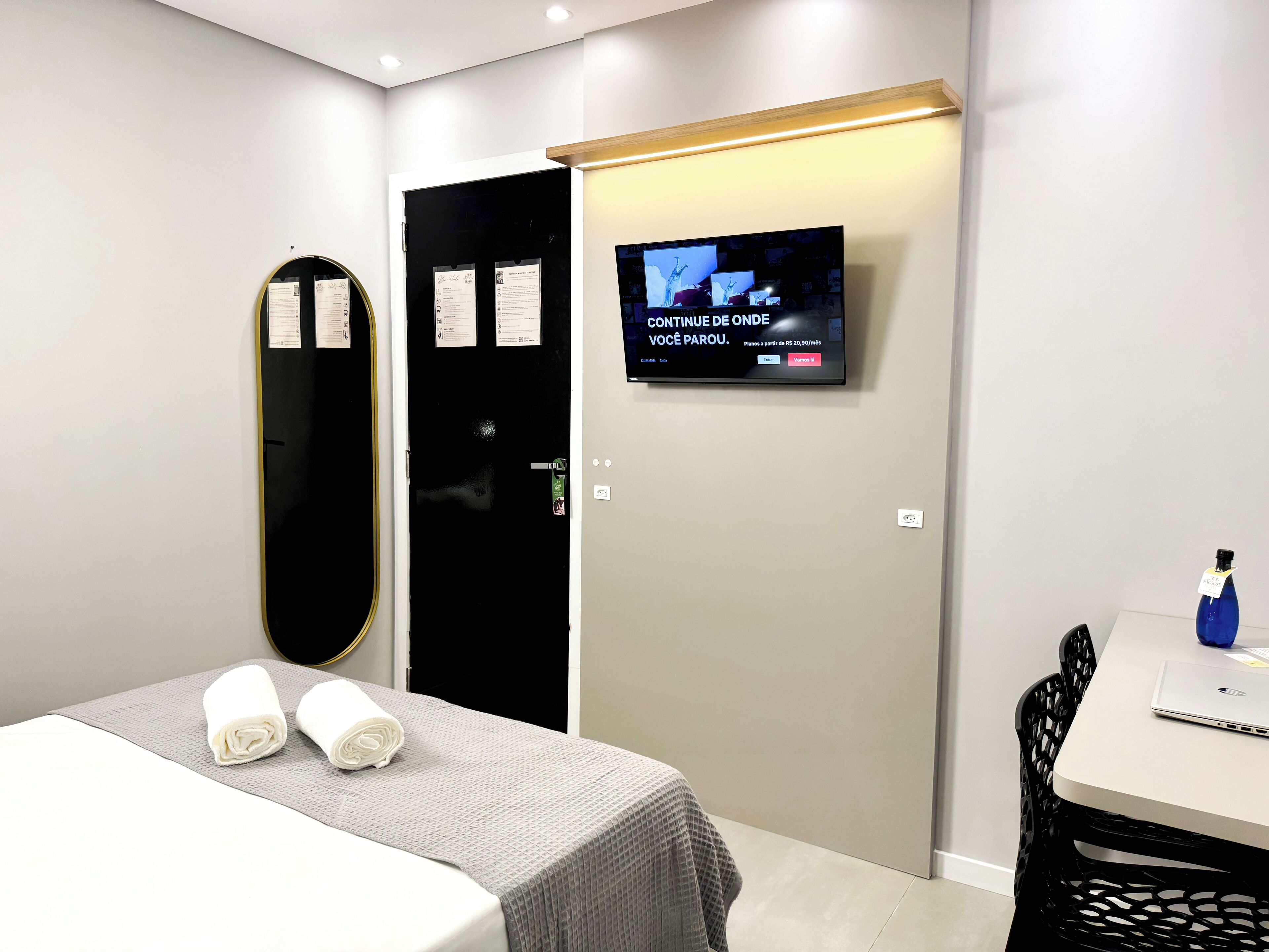 Una camera d'albergo con un letto, una scrivania con un portatile, una TV a muro e una porta con uno specchio e un cartello.