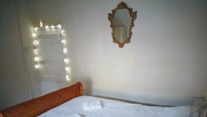 Ein ordentlich gemachtes Bett mit weißen Leinen und einem blauen Kopfkissen, ein hölzernes Kopfteil, ein Spiegel mit aufwendigem Rahmen und Lichterketten an der Wand.