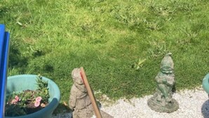 Ein Garten mit einer Statue einer Gestalt, die eine Stange hält, einer hölzernen Bank und einem Blumentopf.