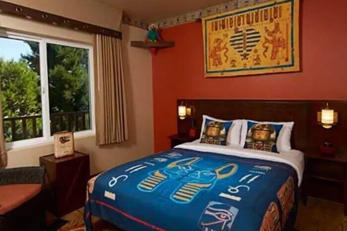 Una habitación de hotel con una cama grande, una ventana con cortinas, un mural en la pared y una mesita de noche con lámpara.