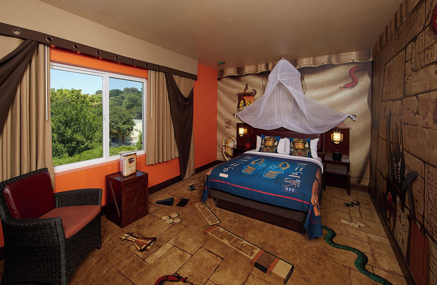 A bedroom with a bed, a chair, a nightstand, a window with curtains, and a wall with Egyptian-themed decorations.
