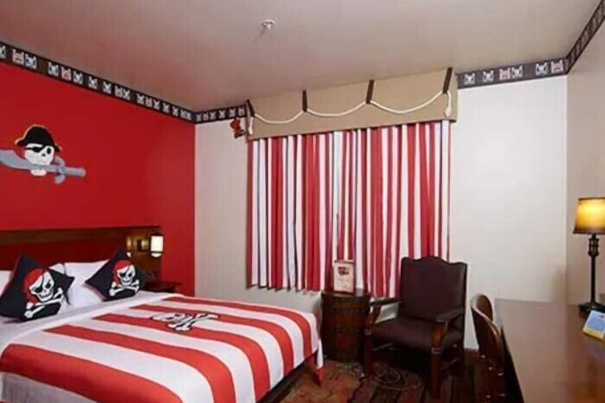 Habitación de hotel con cama con colcha a rayas, cabecera de madera, mesita de noche, silla, escritorio y ventana con persianas verticales.
