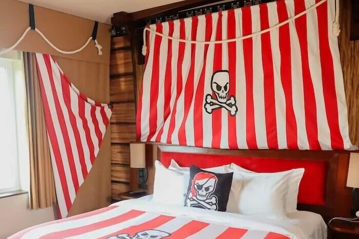 Una habitación con una cama decorada con ropa de cama a rayas rojas y blancas, una almohada con calavera y tibias cruzadas, y una cortina a rayas.