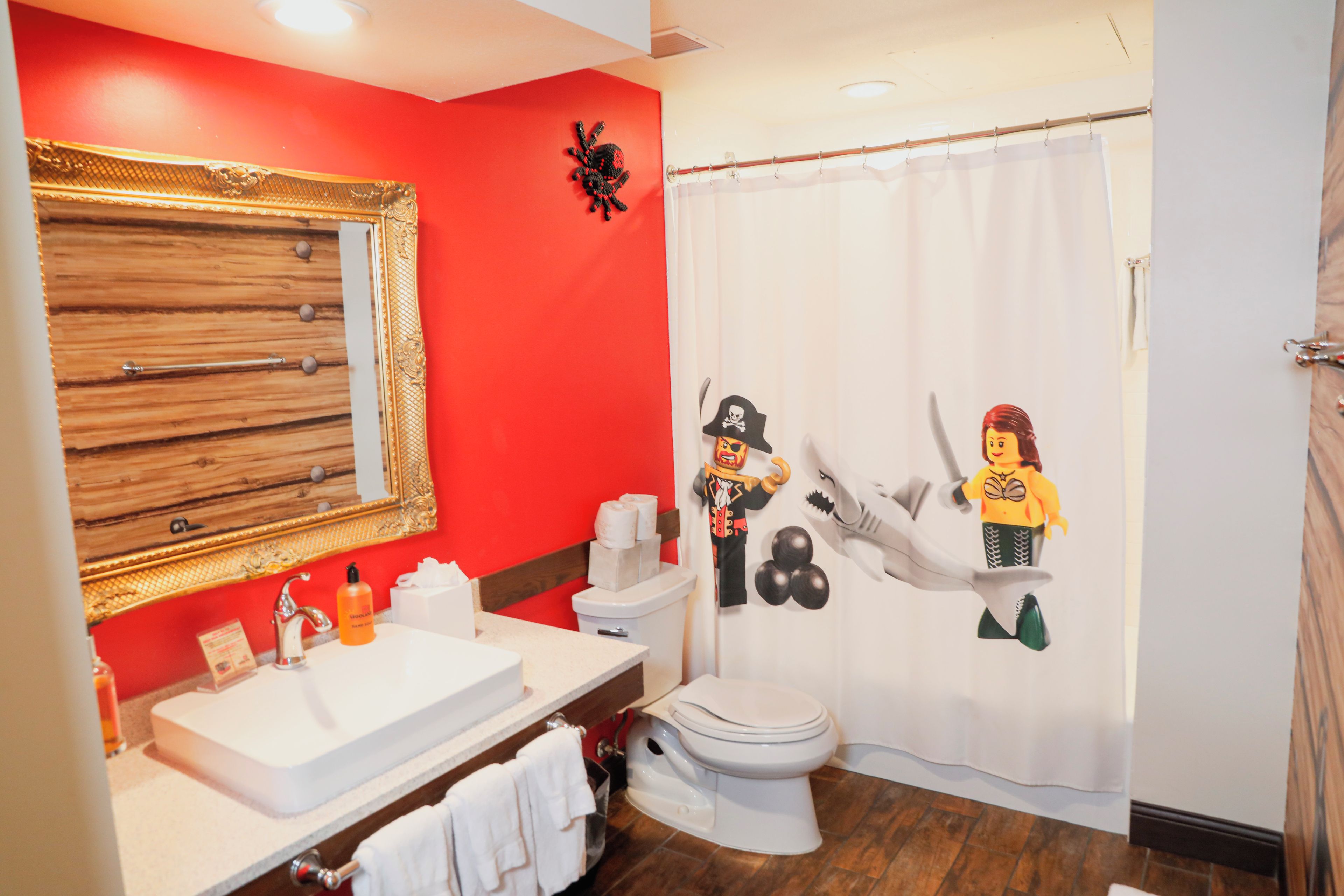 A bathroom with a red wall, a white sink, a toilet, and a shower curtain with a pirate and mermaid design.