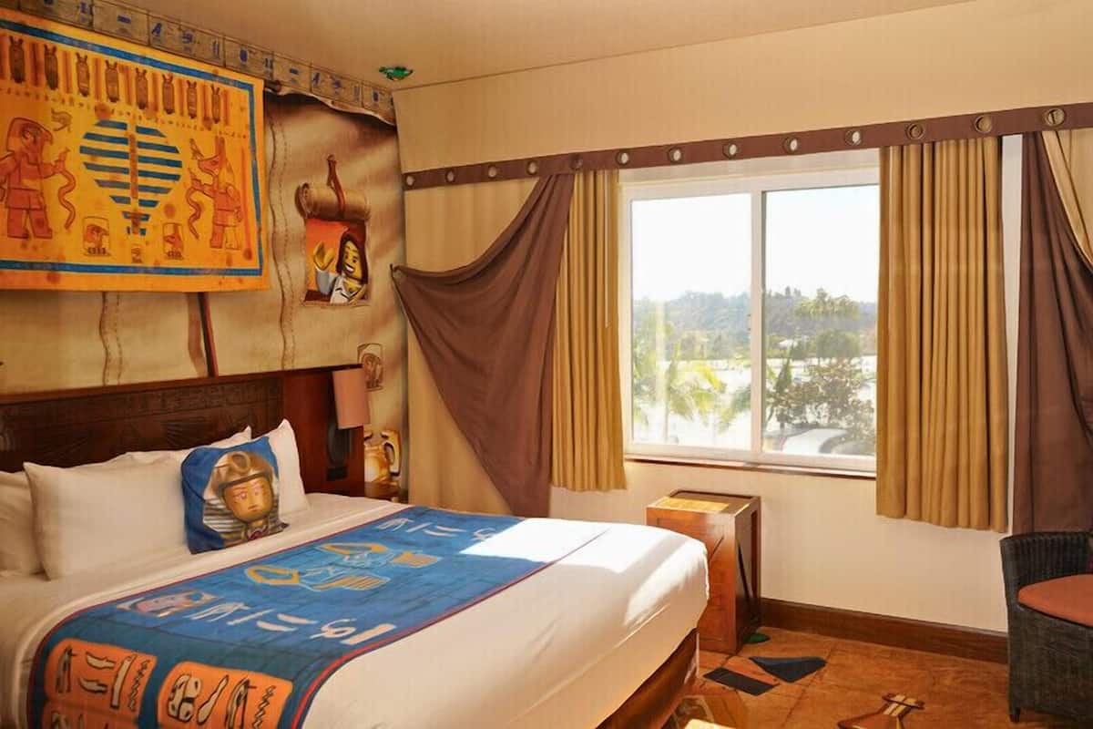 Habitación de hotel con una cama grande, una ventana con cortinas, un cabecero decorado con motivos egipcios y vistas al exterior.