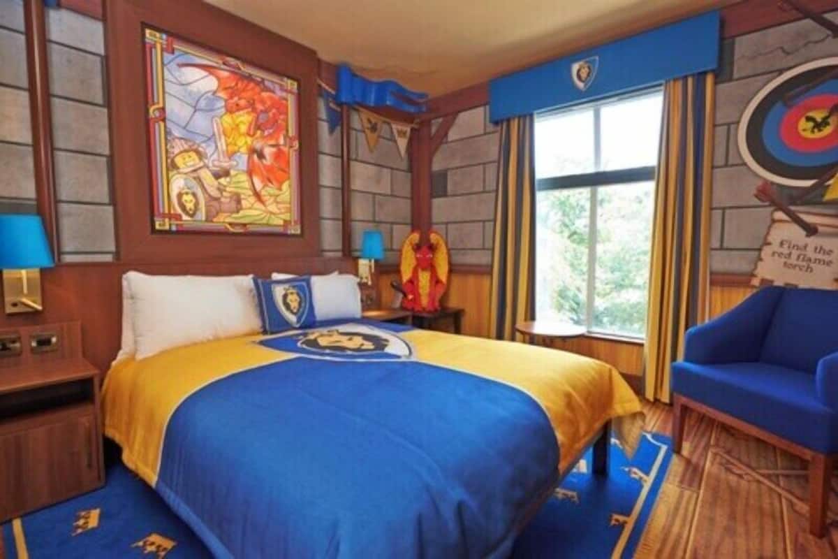 Una habitación con una cama, un sillón azul, un cartel con un blanco y un vitral.