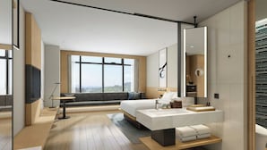 A modern hotel room with a large bed, a sitting area with a sofa and a coffee table, a bathroom with a large mirror, and a view of the city.