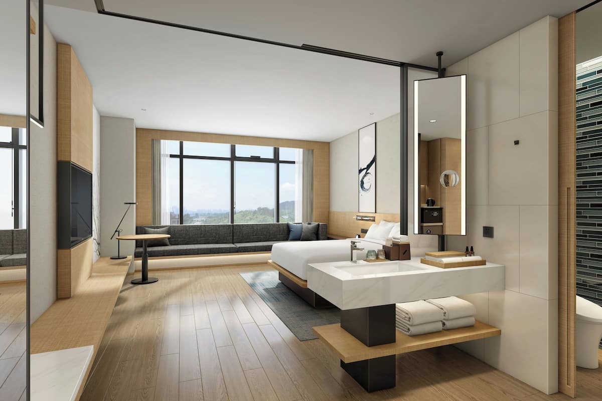 A modern hotel room with a large bed, a sitting area with a sofa and a coffee table, a bathroom with a large mirror, and a view of the city.