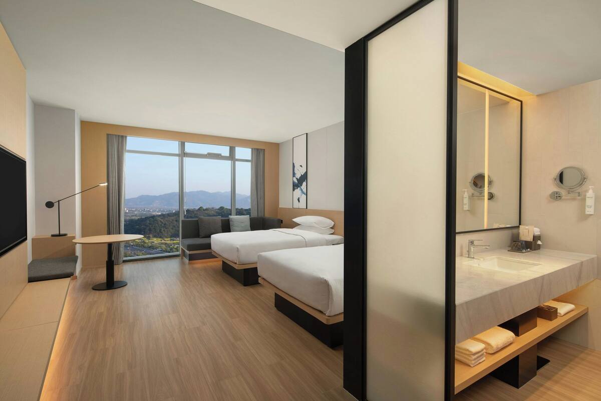 A modern hotel room with two beds, a large window with a city view, a large mirror, and a bathroom with a sink and mirror.