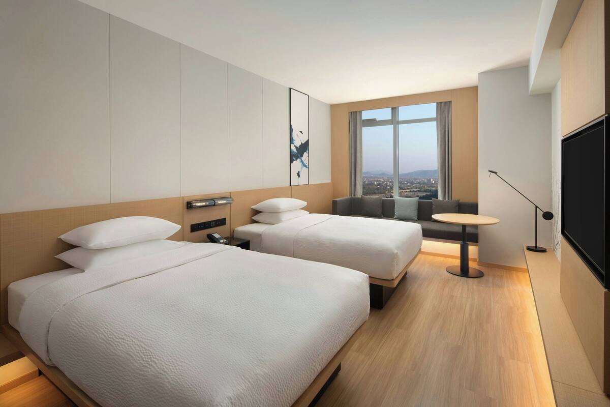 A hotel room with two beds, a small table, a sofa, a TV, and a window with a city view.