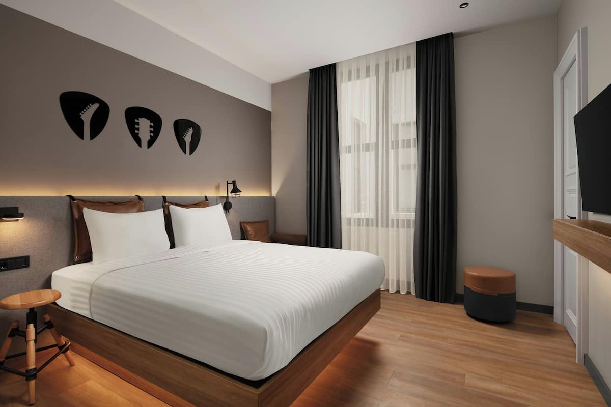 A modern hotel room with a large bed, a wooden floor, a small round ottoman, and a TV on the right side.