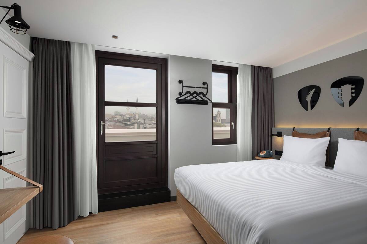 A hotel room with a large bed, a desk, a window with curtains, and a view of the cityscape.
