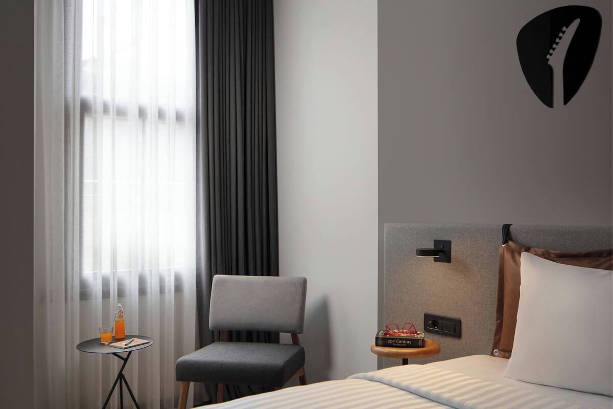 A modern hotel room with a bed, a chair, a small table with a drink and a book, and a wall-mounted light.