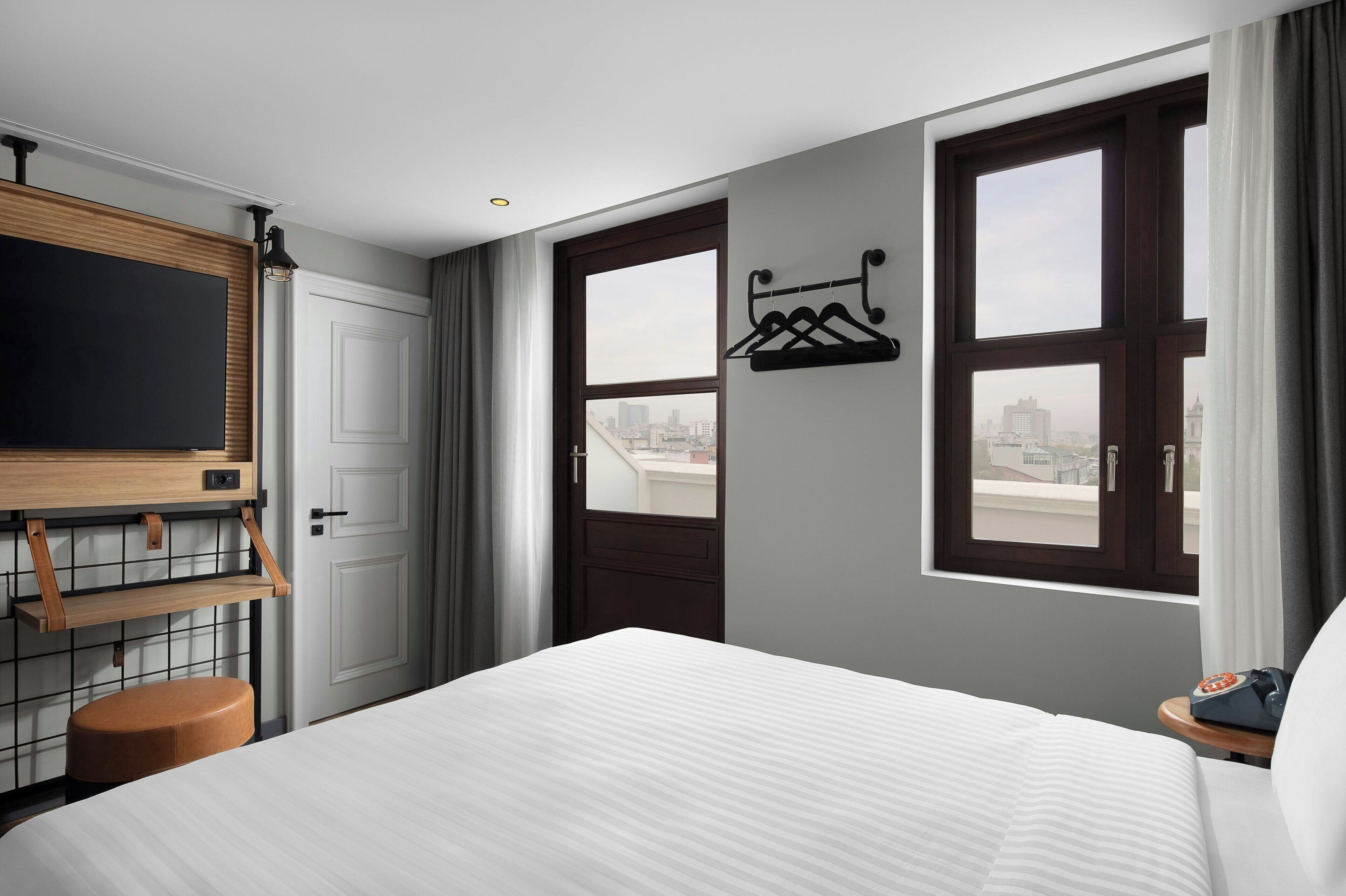 A hotel room with a bed, a TV, a coat rack, and a city view through the windows.