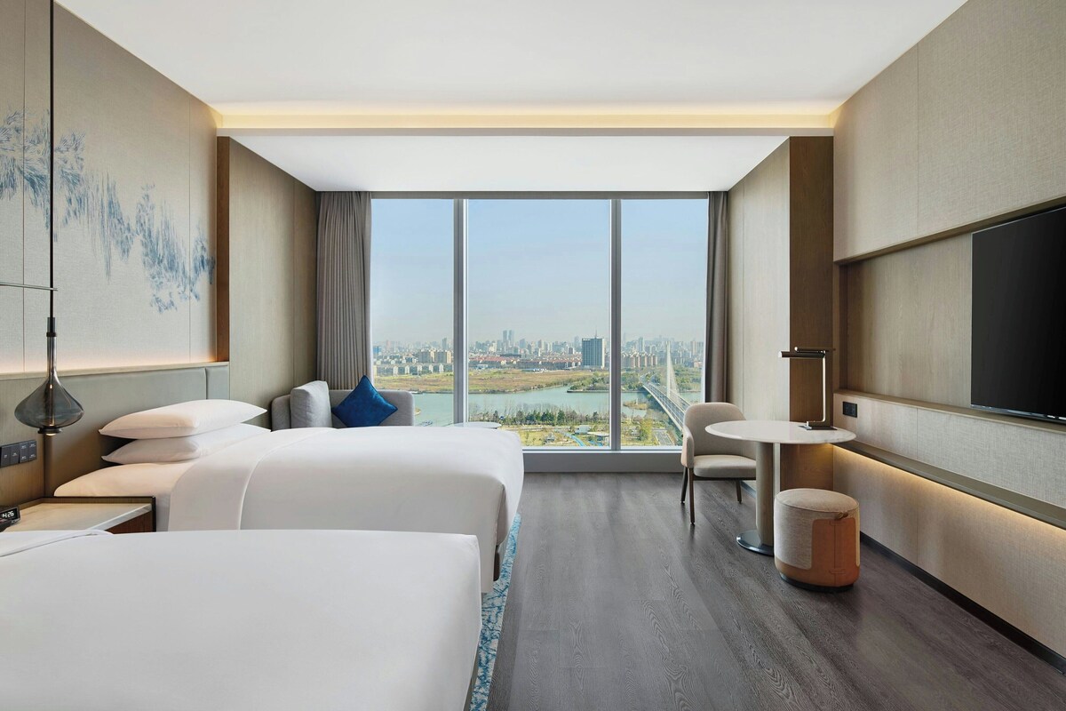 A modern hotel room with two beds, a city view, a round table, and a TV.