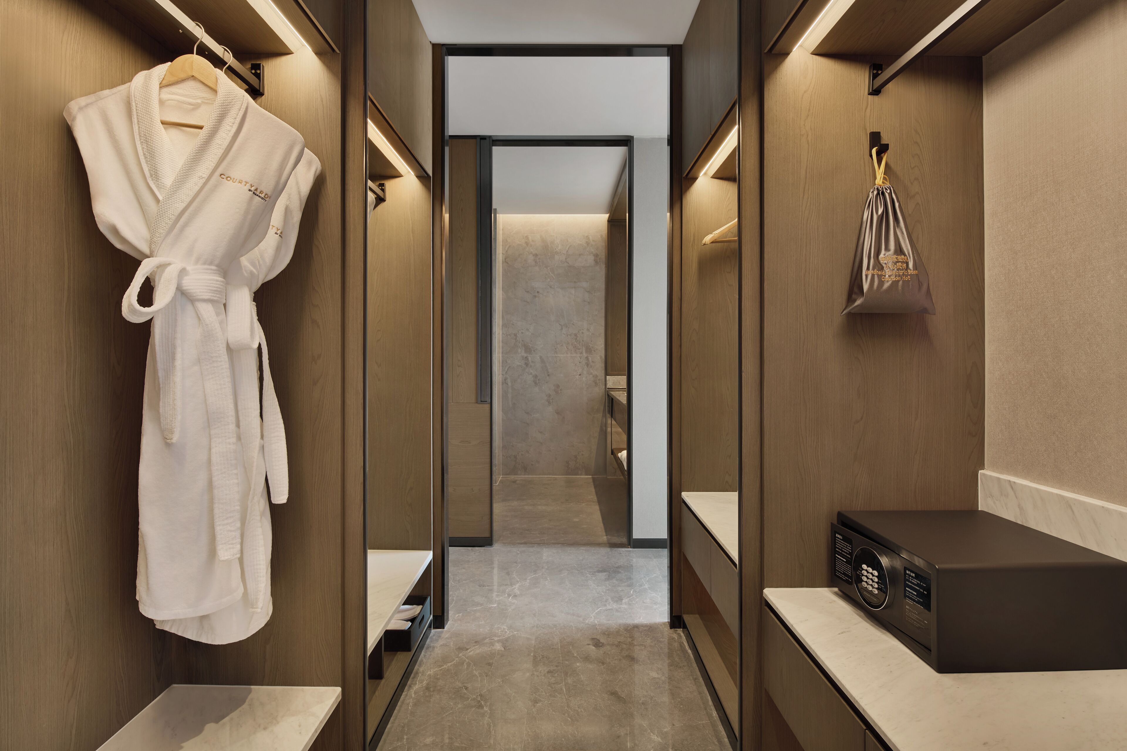 Un baño de hotel moderno con una bata colgada a la izquierda, un perchero a la derecha y un electrodoméstico negro sobre el mostrador.