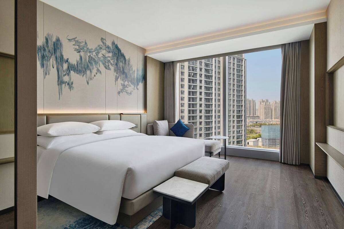 Une chambre d’hôtel moderne avec un grand lit, une petite table, un banc et une vue sur des immeubles de grande hauteur.