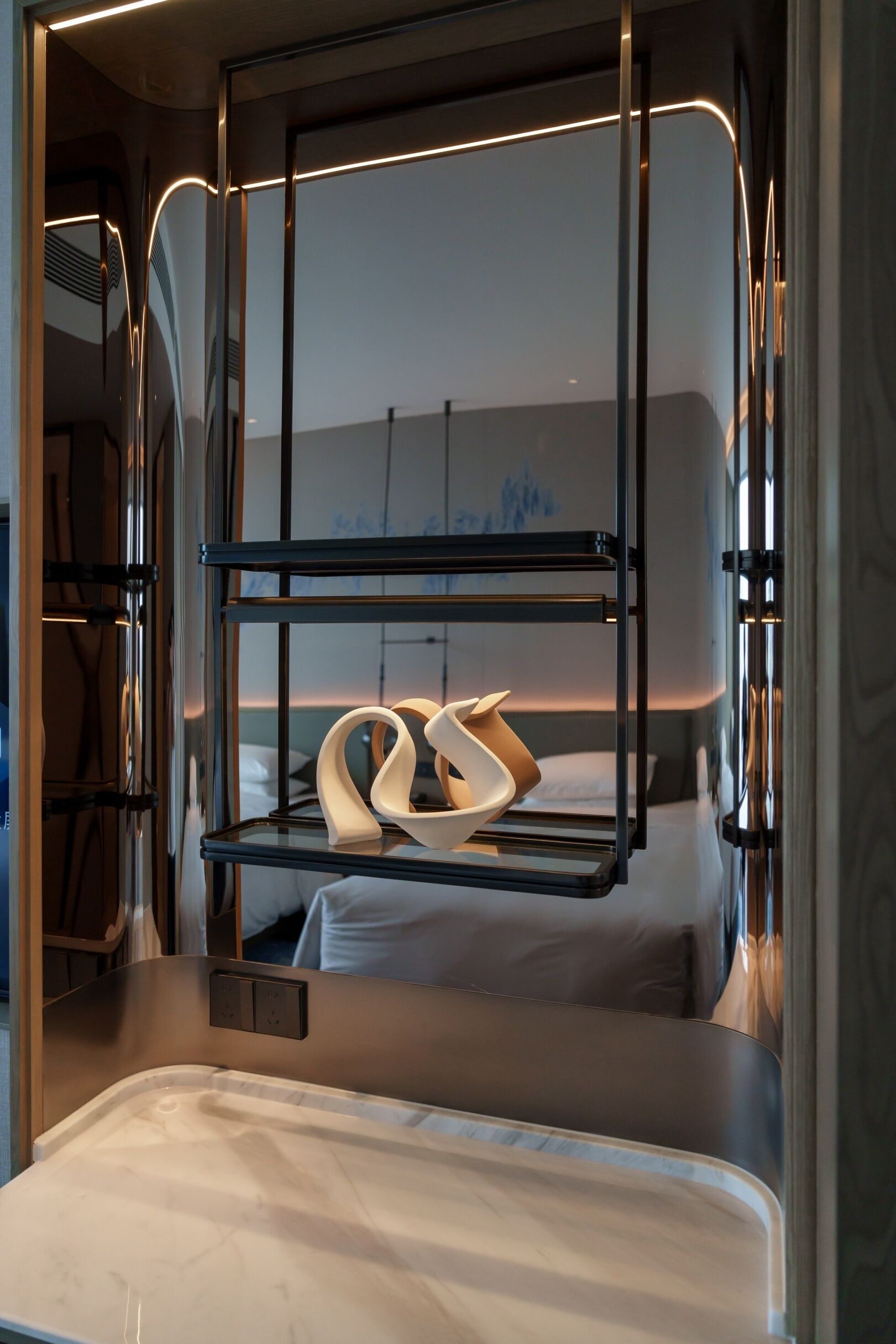 Une chambre d’hôtel moderne avec une cloison en verre, un lit avec une literie blanche et une étagère intégrée où sont posées une paire de pantoufles blanches.