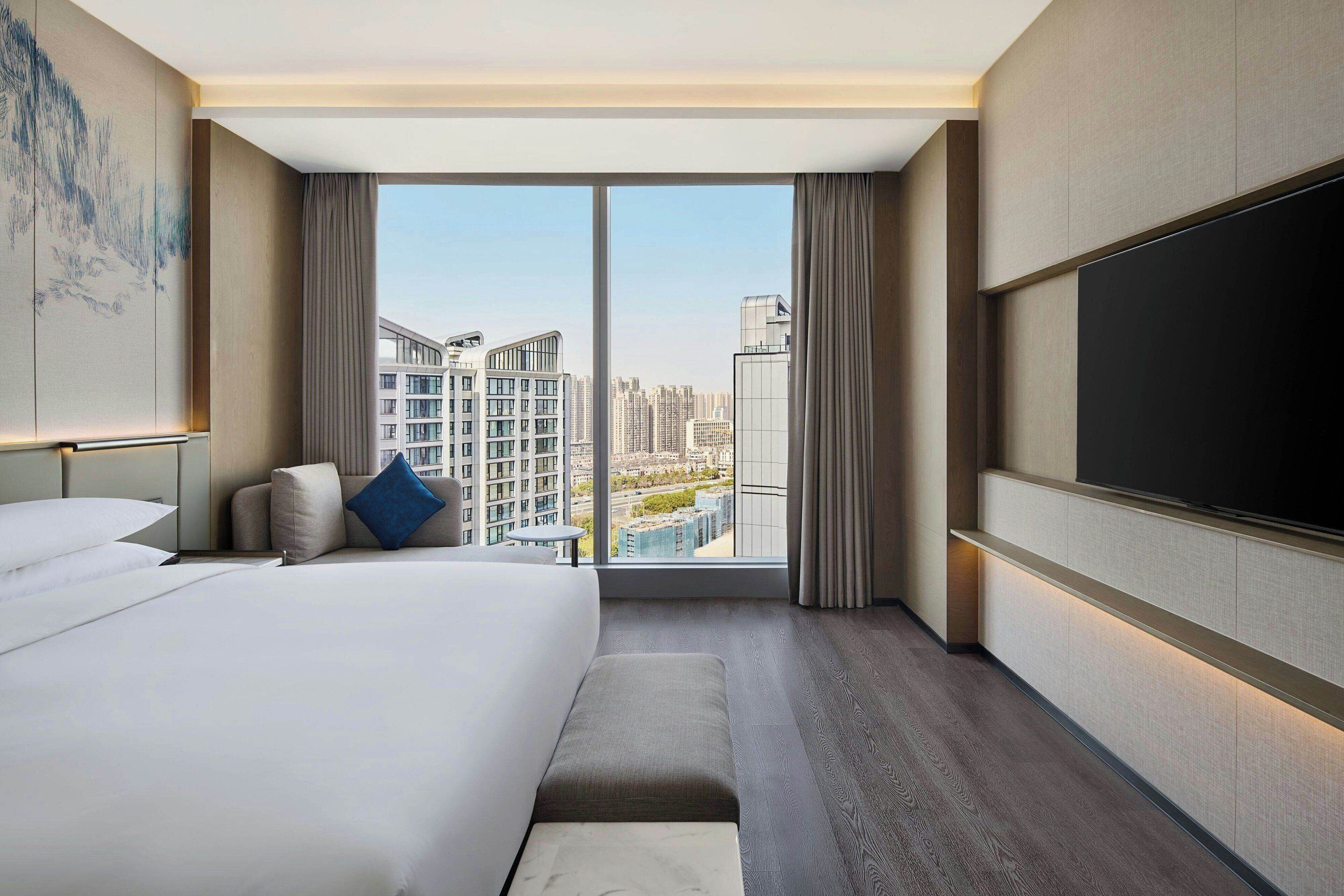 A modern hotel room with a large bed, a flat-screen TV, a sofa, and a view of the cityscape through large windows.