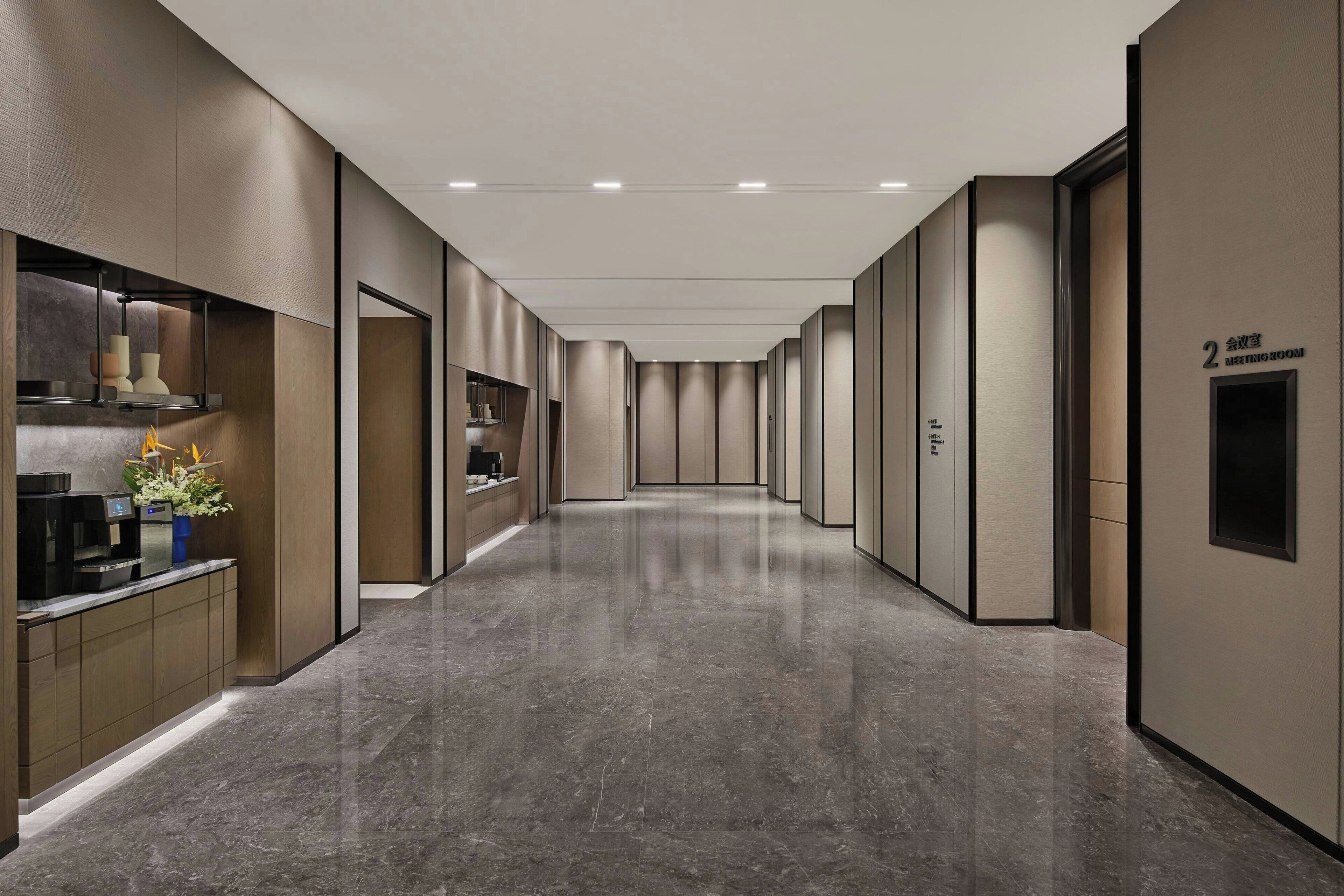 A modern hotel corridor with a sleek design, featuring a reception area with a coffee machine and decorative plants, and a sign indicating the meeting room.