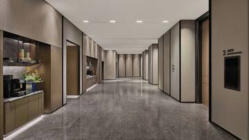 A modern hotel corridor with a sleek design, featuring a reception area with a coffee machine and decorative plants, and a sign indicating the meeting room.