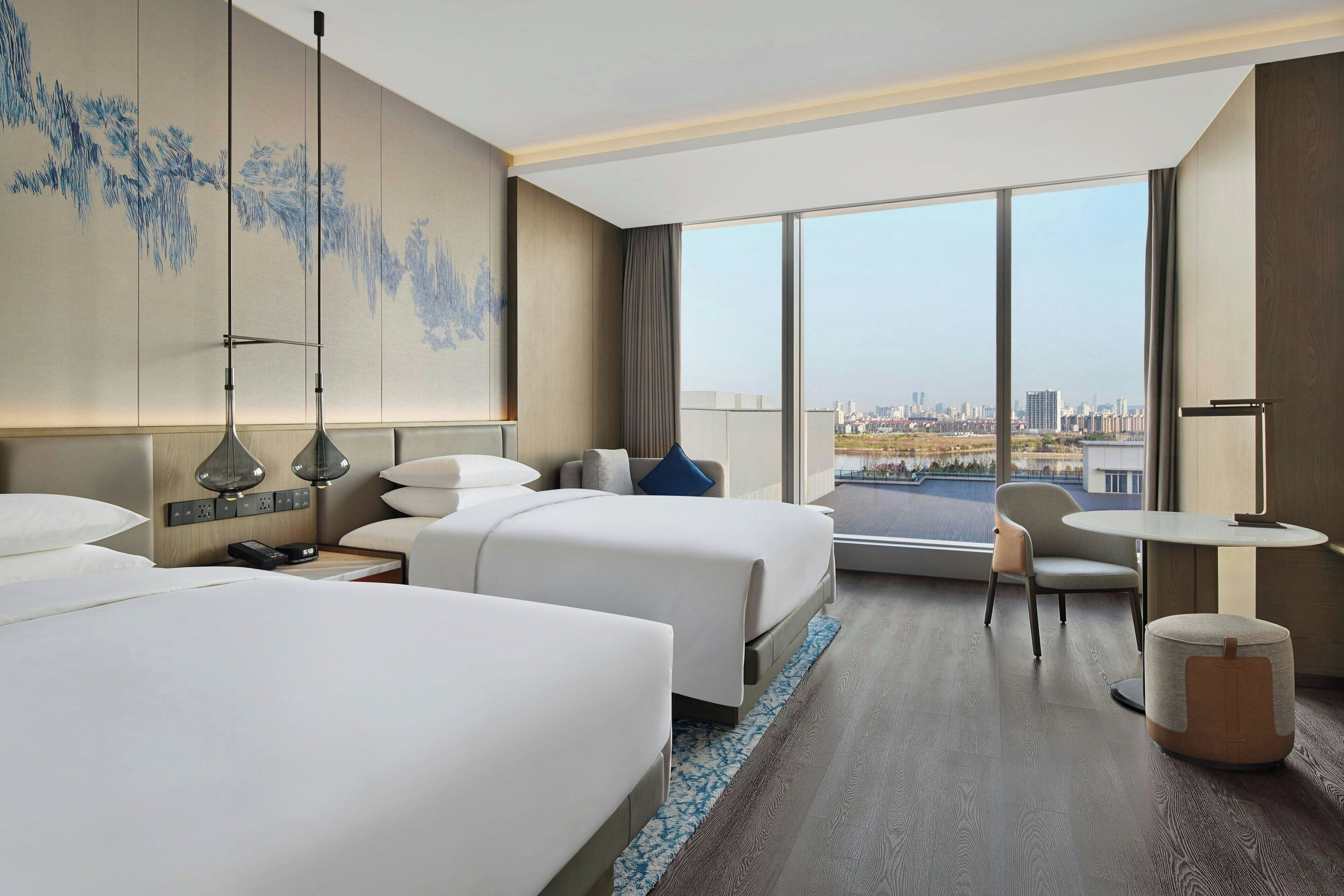 Habitación de hotel con dos camas, un escritorio, una silla y un amplio ventanal con vista a la ciudad.
