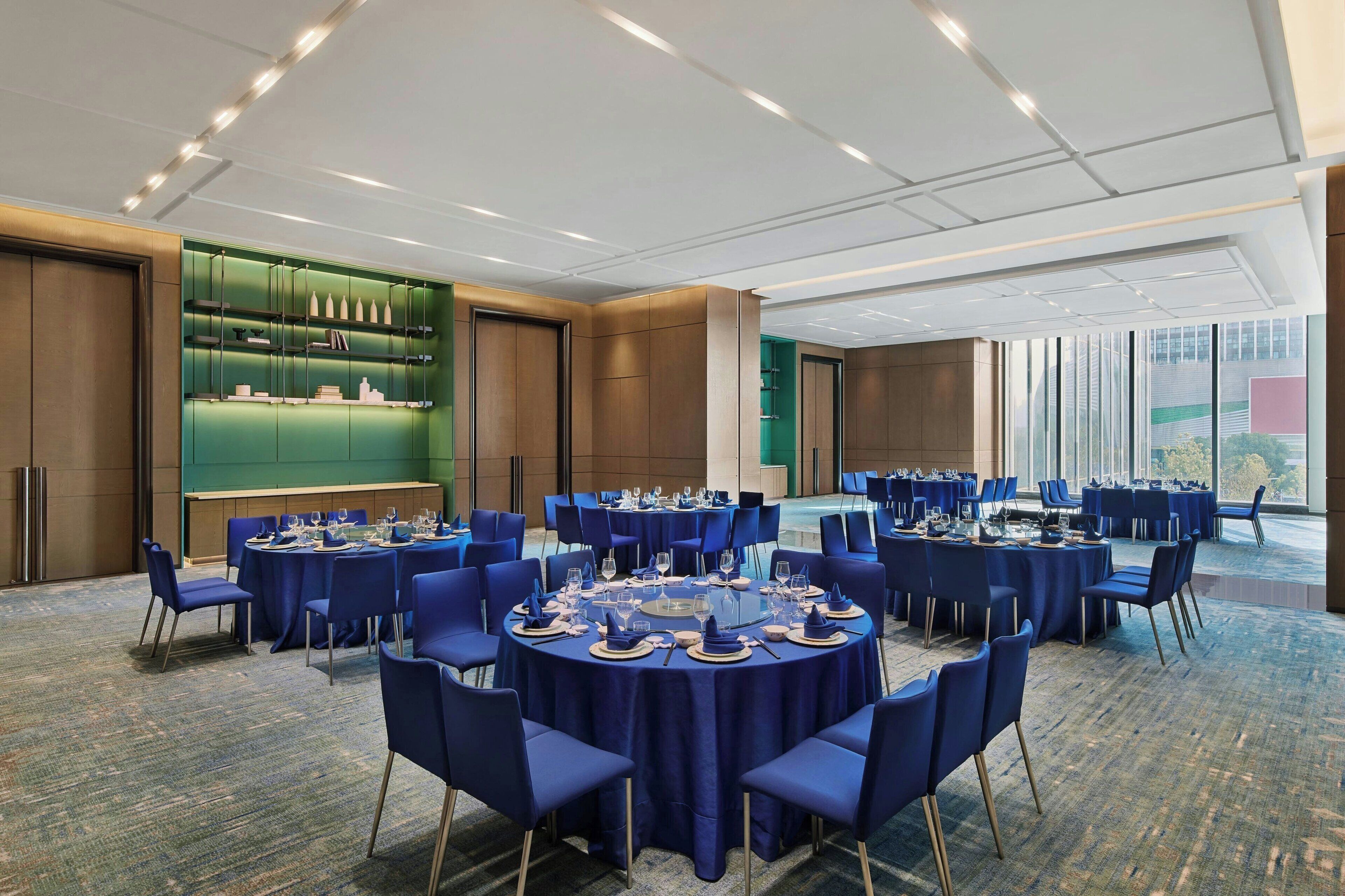 Un salón de conferencias con mesas redondas, sillas azules y una pared de visualización verde.