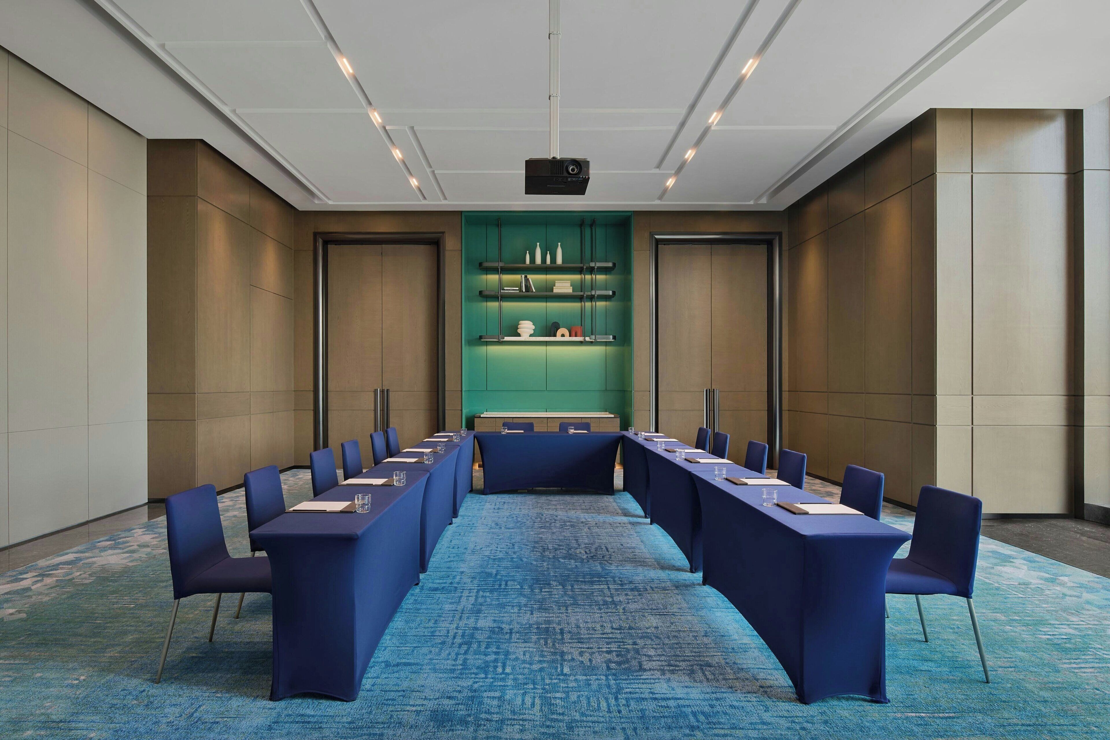Un salón de conferencias con una larga mesa, sillas azules, un proyector y una pared verde con estanterías.