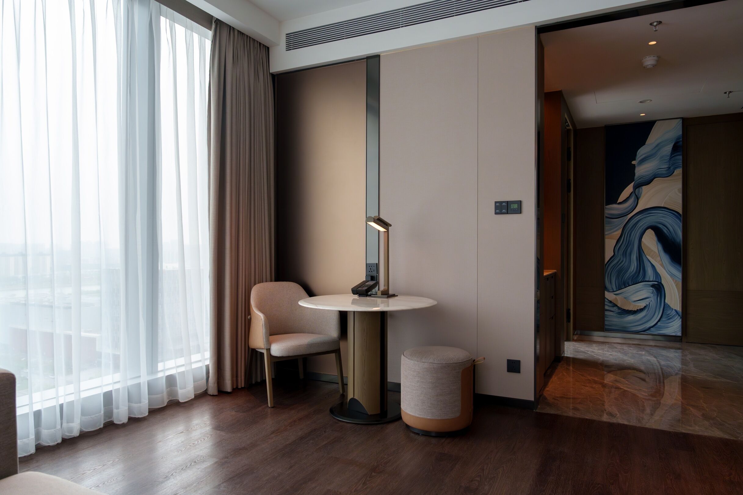 Una habitación de hotel moderna con una mesa redonda, una silla, un pequeño otoman y un ventanal con cortinas ligeras.