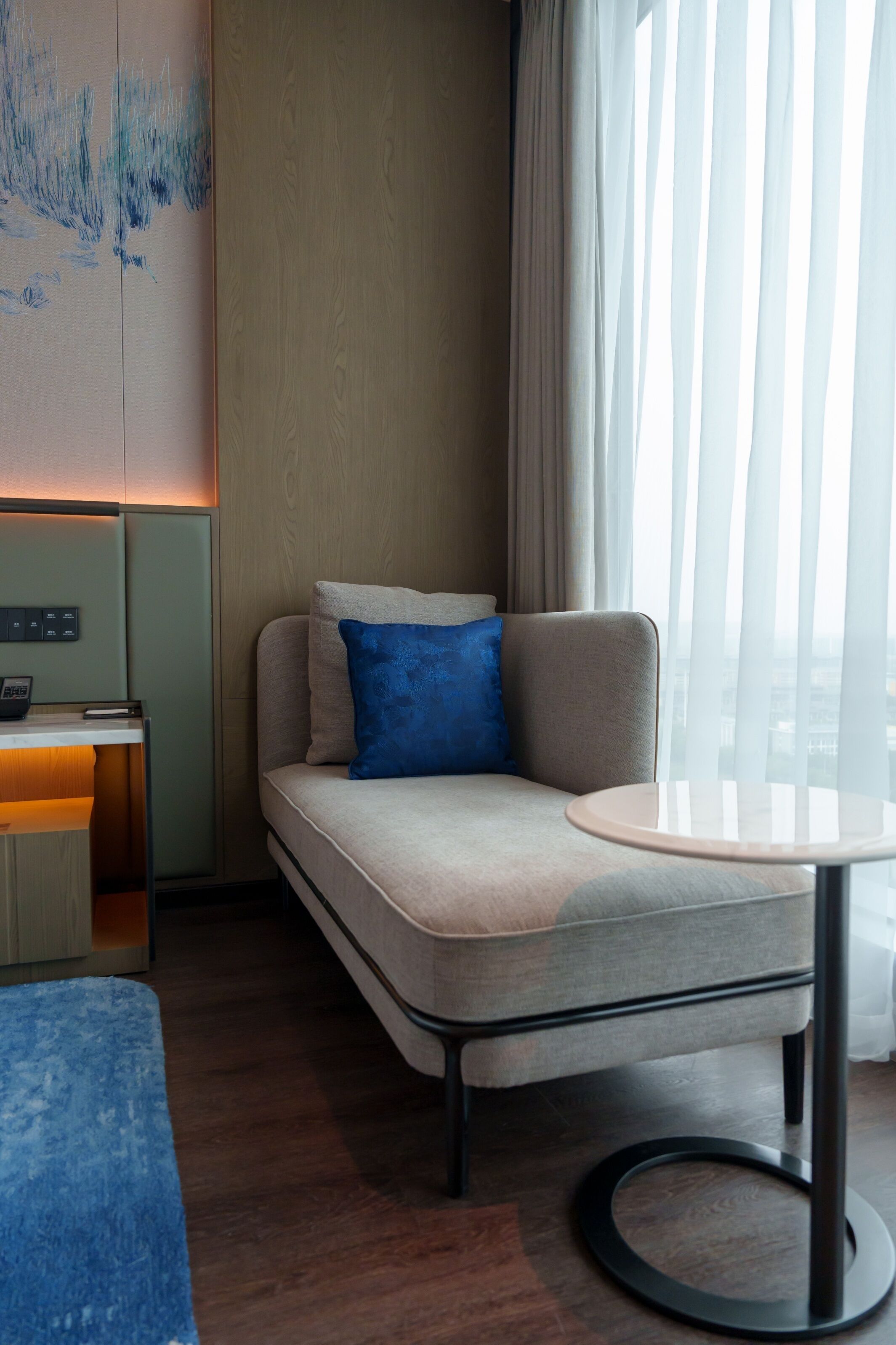 A modern hotel room with a sofa, a small table, a desk, and a view of the city through sheer curtains.
