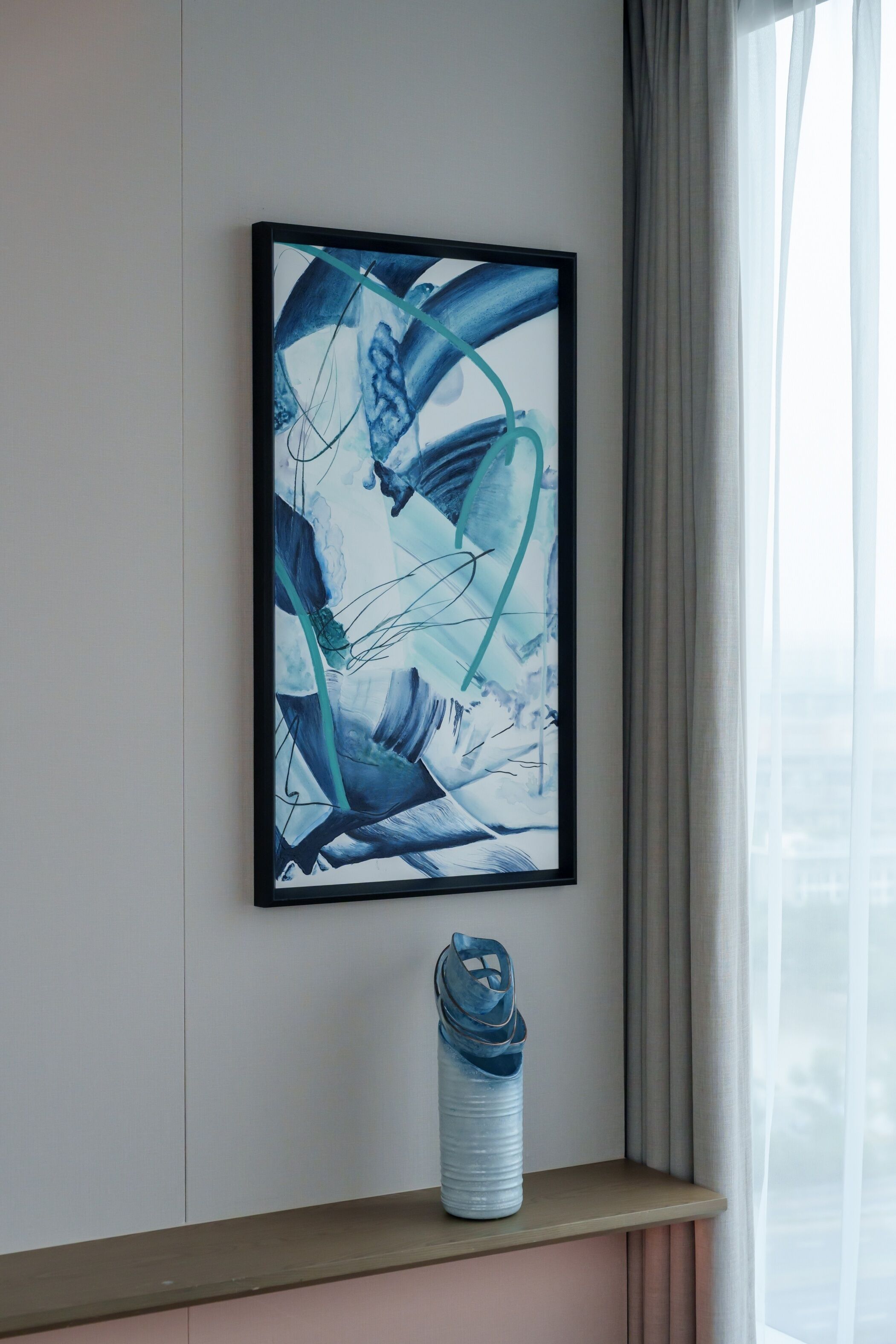 Un cuadro abstracto enmarcado en una pared, una ventana con cortinas y un estante pequeño con una botella de agua.