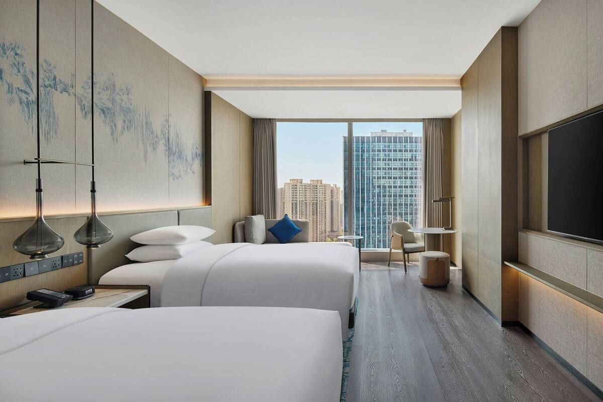 Une chambre d’hôtel avec deux lits, un bureau, une chaise, une télévision et une grande fenêtre donnant sur la ville.