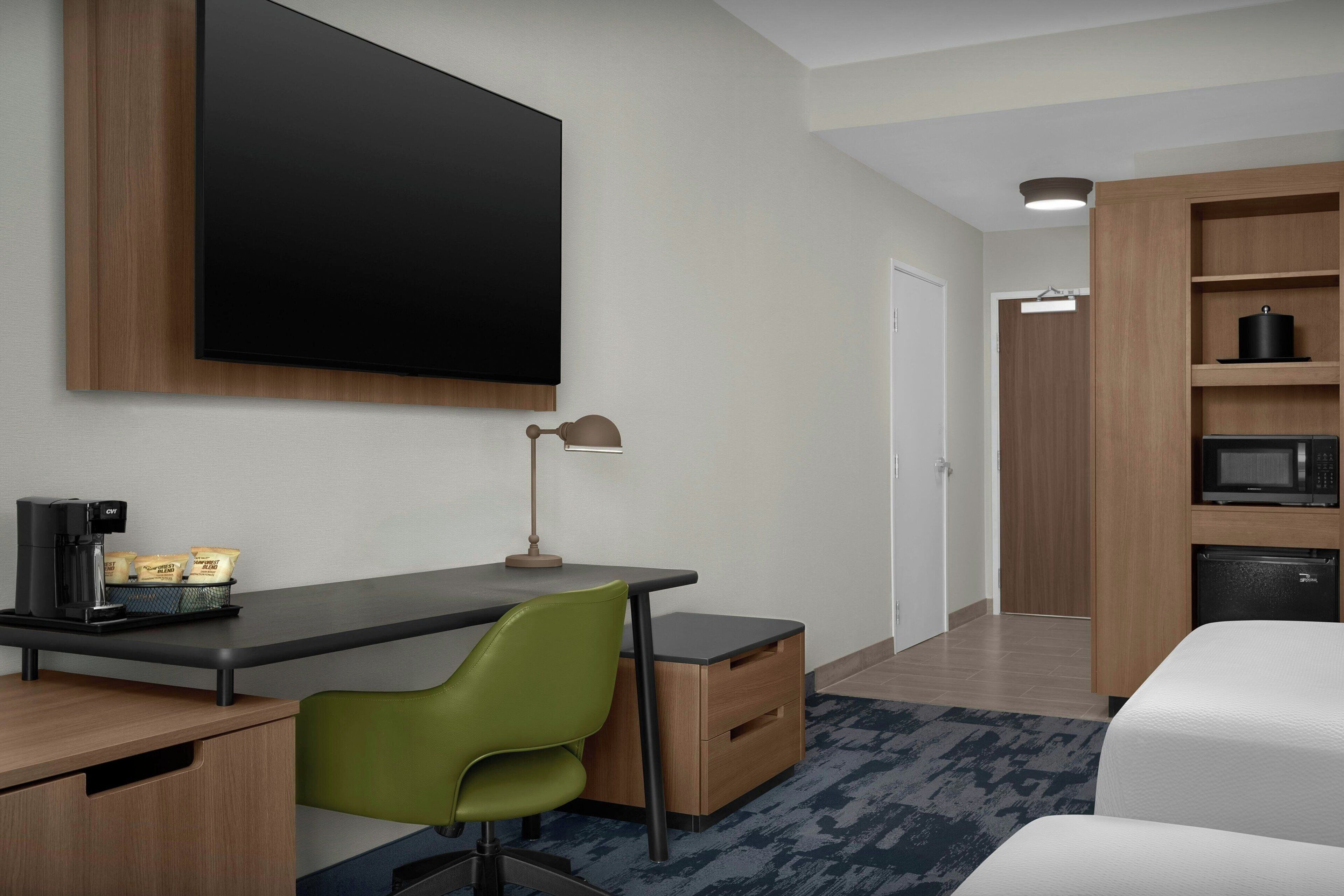 A modern hotel room with a flat-screen TV, a desk with a coffee maker, a green chair, a bed, a wardrobe, and a microwave.