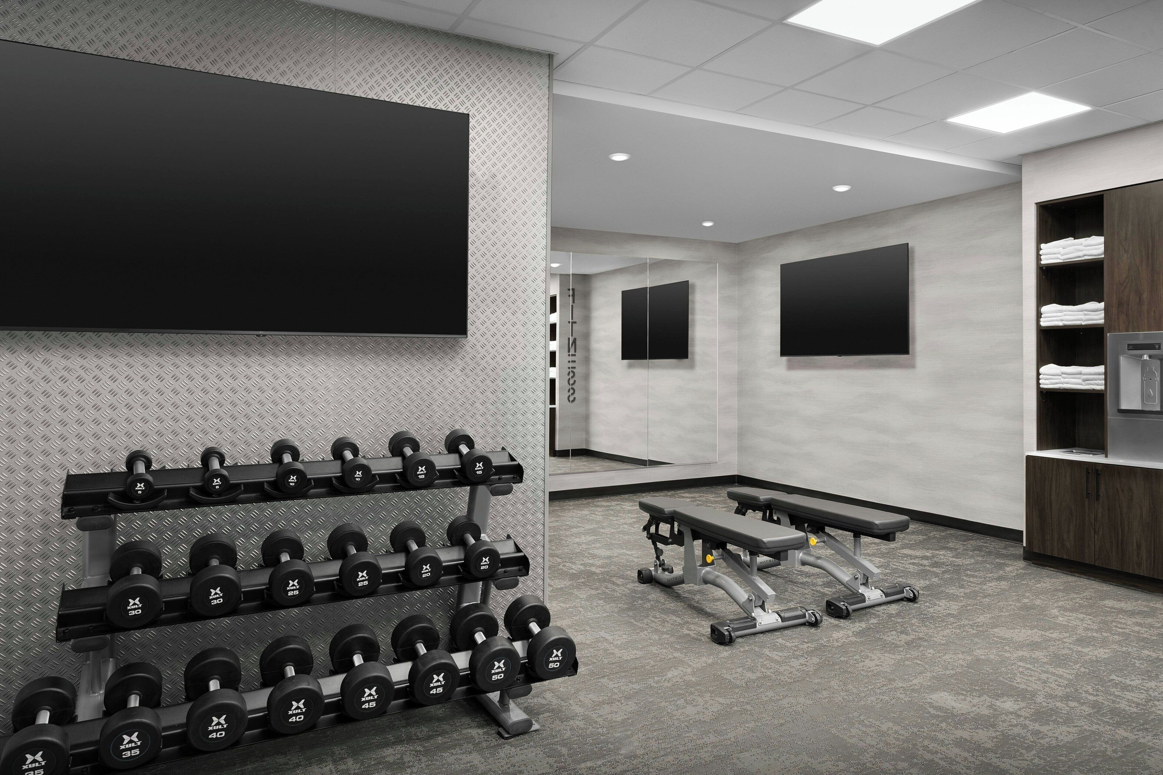 Une salle de sport moderne équipée de supports pour les poids, de bancs et d’un grand écran de télévision.