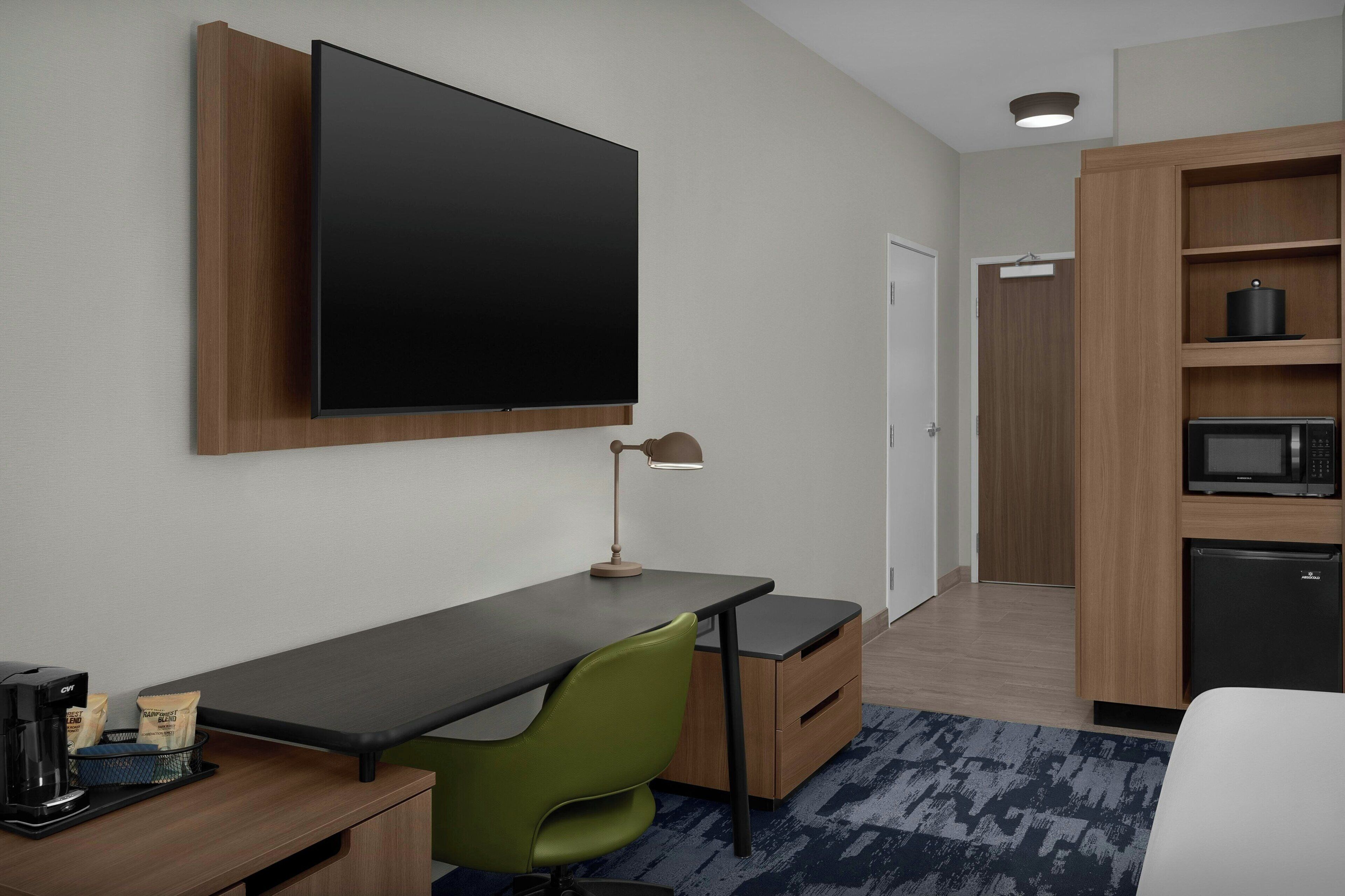 Habitación de hotel con televisor de pantalla plana montado en la pared, escritorio con lámpara, una silla verde, cafetera pequeña y microondas.