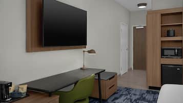 Habitación de hotel con televisor de pantalla plana montado en la pared, escritorio con lámpara, una silla verde, cafetera pequeña y microondas.