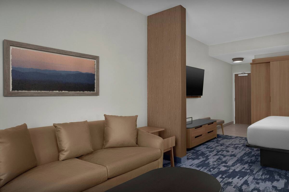 Moderni hotellihuone, jossa on beigenvärinen sohva, seinälle asennettu litteänäyttöinen televisio, puinen vaatekaappi ja sinikuvioinen matto sängyn alla.