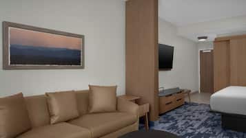 A modern hotel room with a beige sofa, a flat-screen TV mounted on the wall, a wooden wardrobe, and a bed with a blue patterned rug.