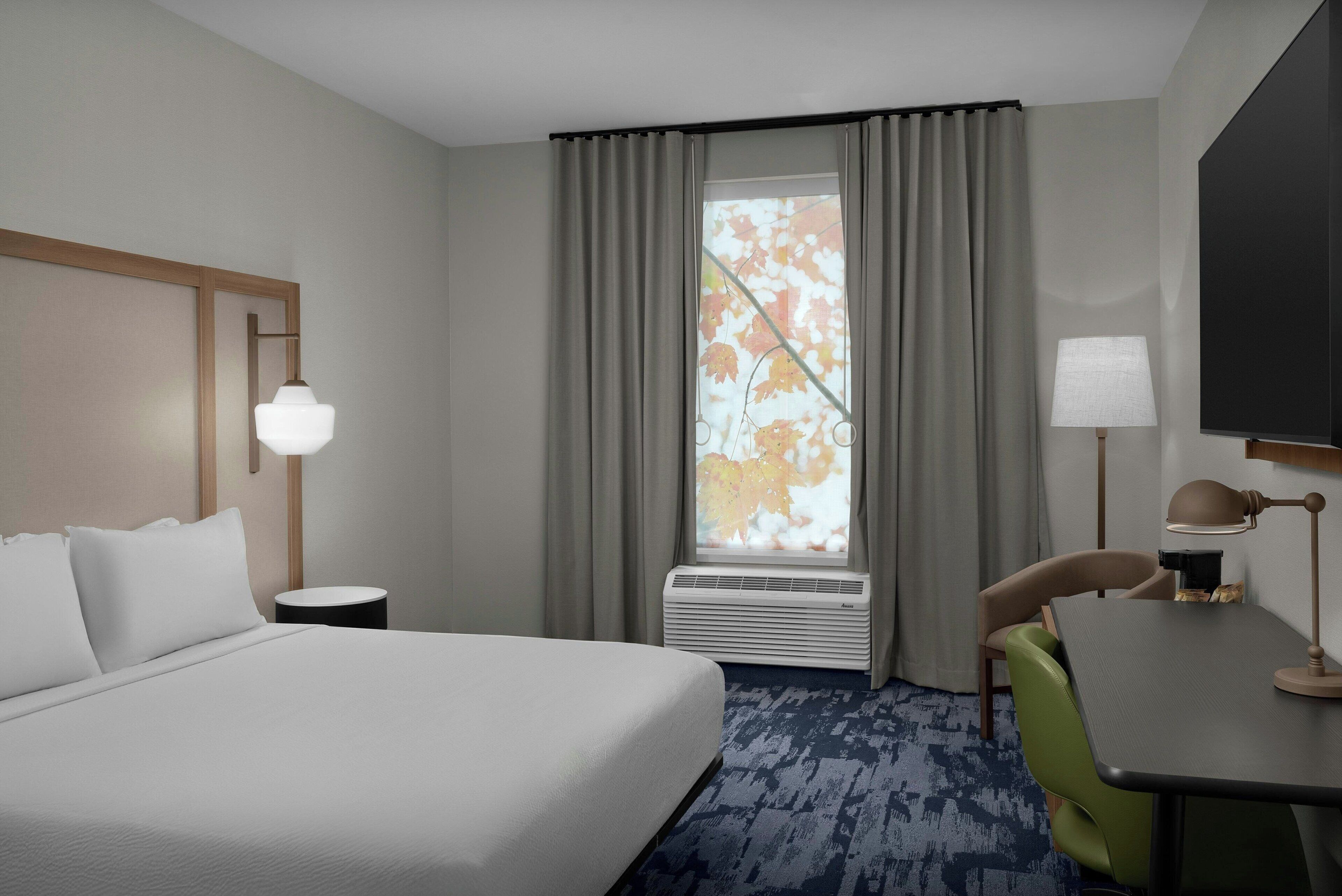 A hotel room with a bed, a desk, a chair, a lamp, a window with curtains, and an air conditioning unit.
