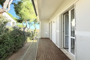 Une terrasse en bois couverte, agrémentée de volets blancs, et entourée de verdure et d’arbres.
