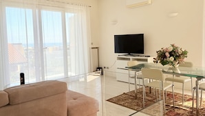 Una sala de estar moderna con un sofá, una mesa de comedor de vidrio, sillas, un televisor y un ventanal con cortinas.