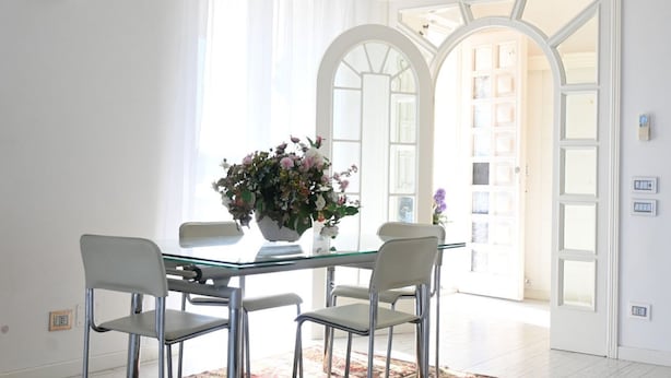 Un comedor con una mesa de vidrio, cuatro sillas, un jarrón de flores y vistas a un interior blanco a través de un arco.