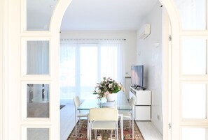 Une porte ouverte donne sur un coin repas, où l’on trouve une table en verre, des chaises et un vase rempli de fleurs.